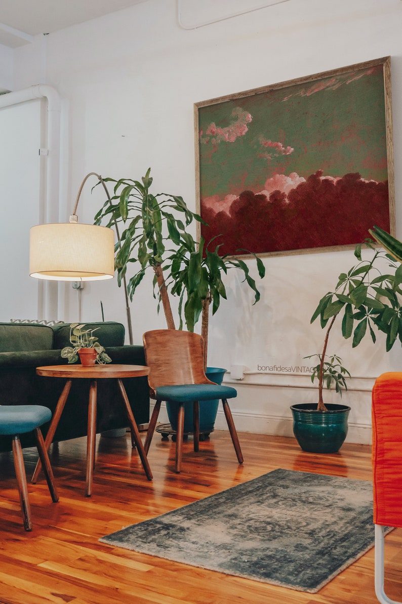 May include: A living room interior with a green and red abstract painting on the wall, a green potted plant, a wooden chair with a blue cushion, a small round wooden table, a green sofa, and a cream-colored lamp.