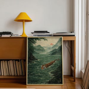 May include: A framed art print depicting a tiger swimming in a river, with mountains and a setting sun. The artwork has a vintage aesthetic, with a green, brown, and orange colour palette. The print is displayed on a wooden table.