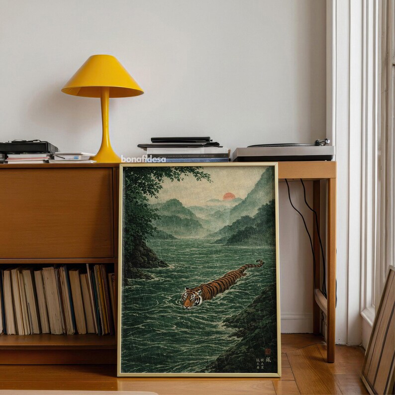May include: A framed art print depicting a tiger swimming in a river, with mountains and a setting sun. The artwork has a vintage aesthetic, with a green, brown, and orange colour palette. The print is displayed on a wooden table.