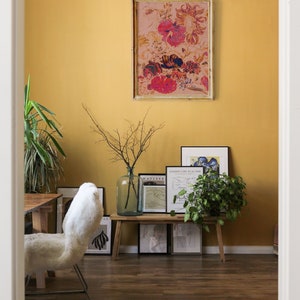 May include: A yellow wall with a framed floral print, a wooden bench with framed prints, a white furry chair, and a potted plant.