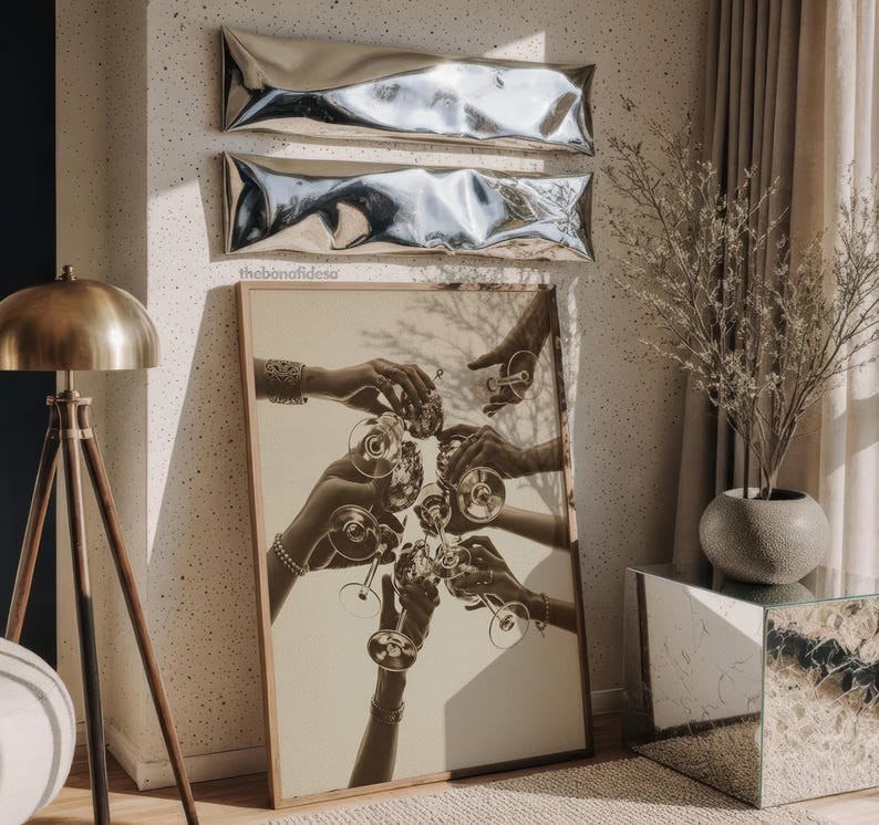 May include: A framed sepia-toned print depicts hands toasting with wine glasses. Above the print are two silver, rectangular wall decorations. A brass floor lamp and a mirrored side table are also visible in the room.