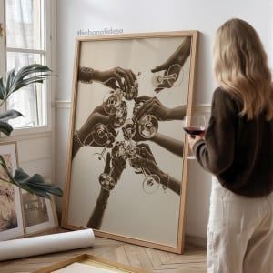 May include: Sepia-toned framed print featuring hands toasting with wine glasses. The image captures a celebratory moment, with multiple hands reaching towards each other. The frame is a light wood color, and the overall aesthetic is vintage.
