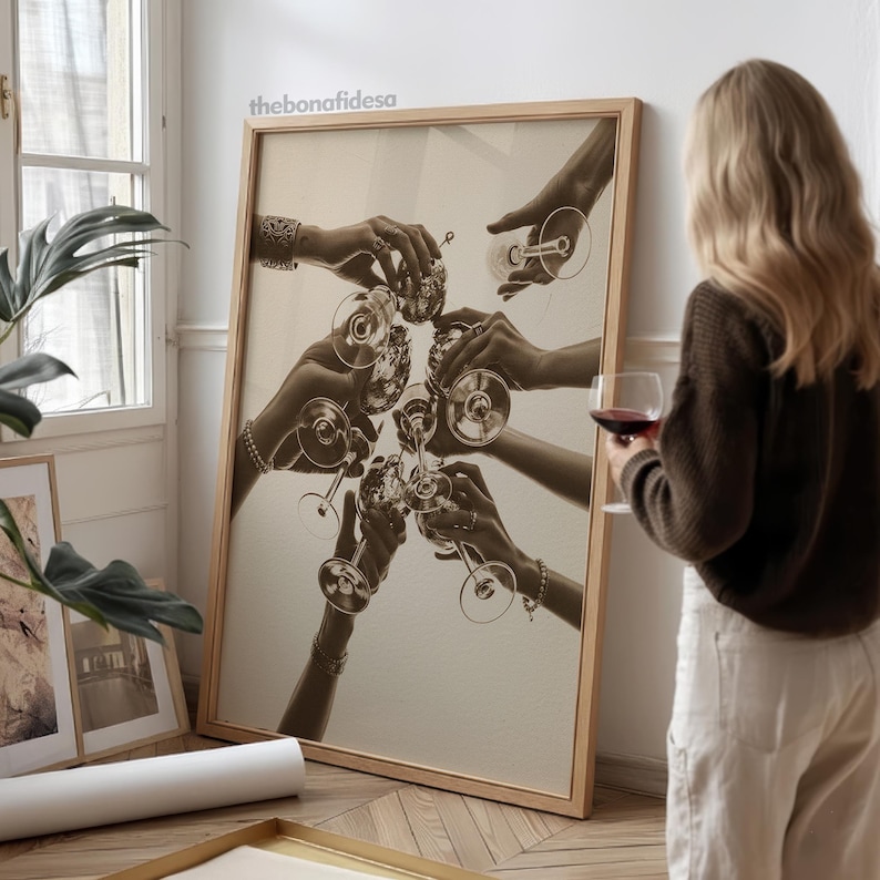 May include: Sepia-toned framed print featuring hands toasting with wine glasses. The image captures a celebratory moment, with multiple hands reaching towards each other. The frame is a light wood color, and the overall aesthetic is vintage.