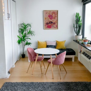 May include: A white round table with four legs and two pink chairs. The table is set with a white tablecloth and a vase of flowers. The chairs are upholstered in a soft pink fabric. The table is set in a living room with a large window and a plant in the corner.