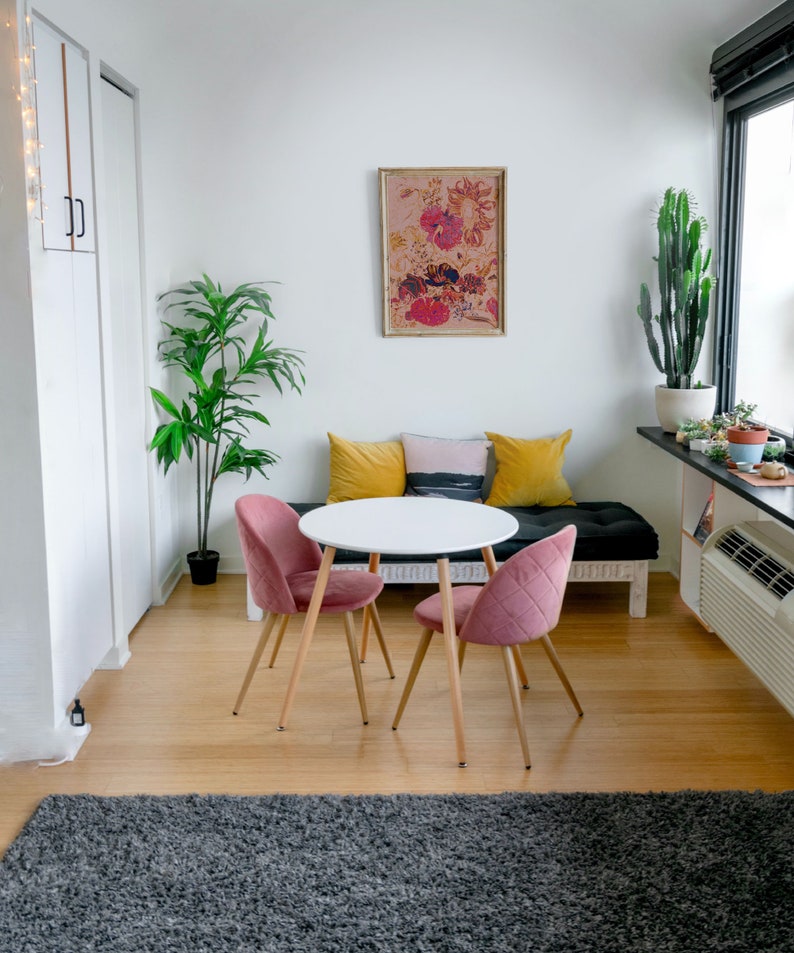 May include: A white round table with four legs and two pink chairs. The table is set with a white tablecloth and a vase of flowers. The chairs are upholstered in a soft pink fabric. The table is set in a living room with a large window and a plant in the corner.