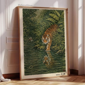 May include: Framed art print featuring a tiger in a lush, green environment. The tiger, with orange and black stripes, is reflected in the water. A dragonfly hovers nearby. The artwork is in a gold frame and leans against a white wall.