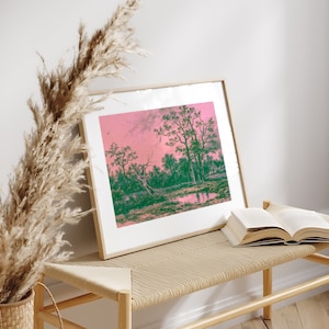 Puede incluir: Una impresi&oacute;n enmarcada de una pintura de paisaje con un cielo rosa y &aacute;rboles verdes reflejados en un cuerpo de agua tranquilo. La impresi&oacute;n est&aacute; sentada en un banco de madera clara con un libro encima.