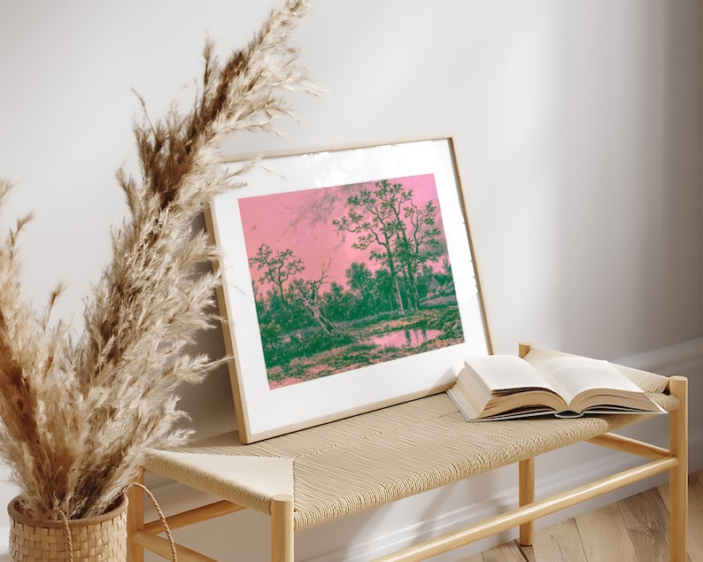 Puede incluir: Una impresi&oacute;n enmarcada de una pintura de paisaje con un cielo rosa y &aacute;rboles verdes reflejados en un cuerpo de agua tranquilo. La impresi&oacute;n est&aacute; sentada en un banco de madera clara con un libro encima.