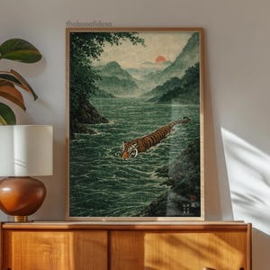 May include: A framed art print depicting a tiger swimming in a river, set against a backdrop of mountains and a setting sun. The artwork has a traditional Japanese style, with a colour palette of greens, browns, and oranges. The print is displayed in a wooden frame.