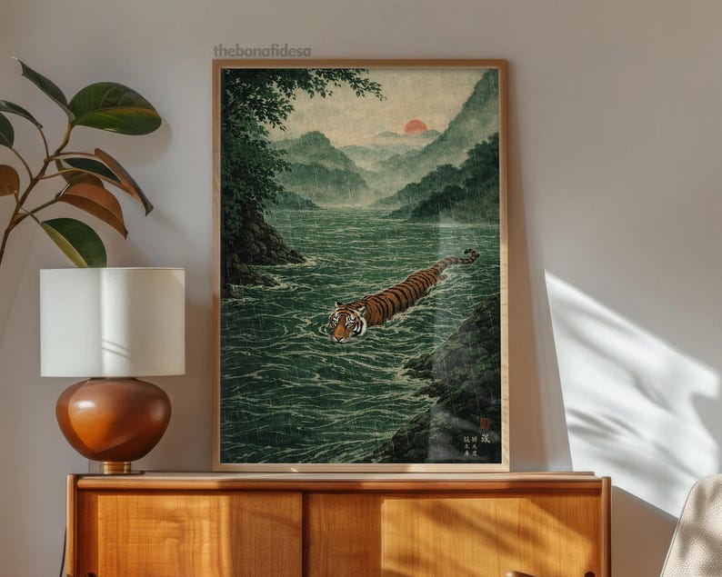 May include: A framed art print depicting a tiger swimming in a river, set against a backdrop of mountains and a setting sun. The artwork has a traditional Japanese style, with a colour palette of greens, browns, and oranges. The print is displayed in a wooden frame.