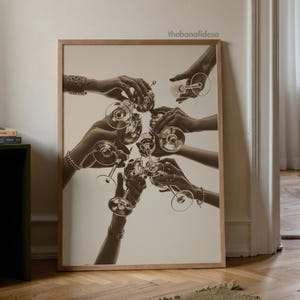 May include: Sepia-toned print of hands toasting with champagne glasses. The image is framed in a light wood frame and features multiple hands reaching towards the center, holding the glasses. The background is a neutral color.
