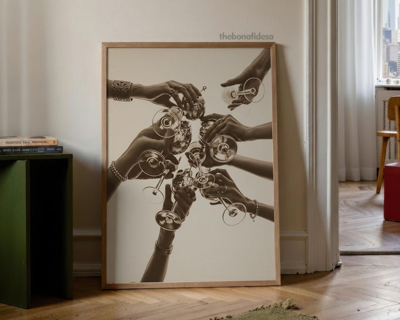 May include: Sepia-toned print of hands toasting with champagne glasses. The image is framed in a light wood frame and features multiple hands reaching towards the center, holding the glasses. The background is a neutral color.
