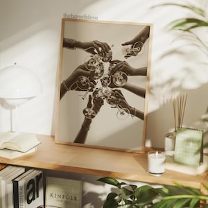 May include: Sepia-toned print of hands toasting with champagne glasses. The image, set in a wooden frame, captures a celebratory moment. The bonafidesa text is visible in the top corner. The print is displayed on a wooden shelf with a lamp, books, and a candle.