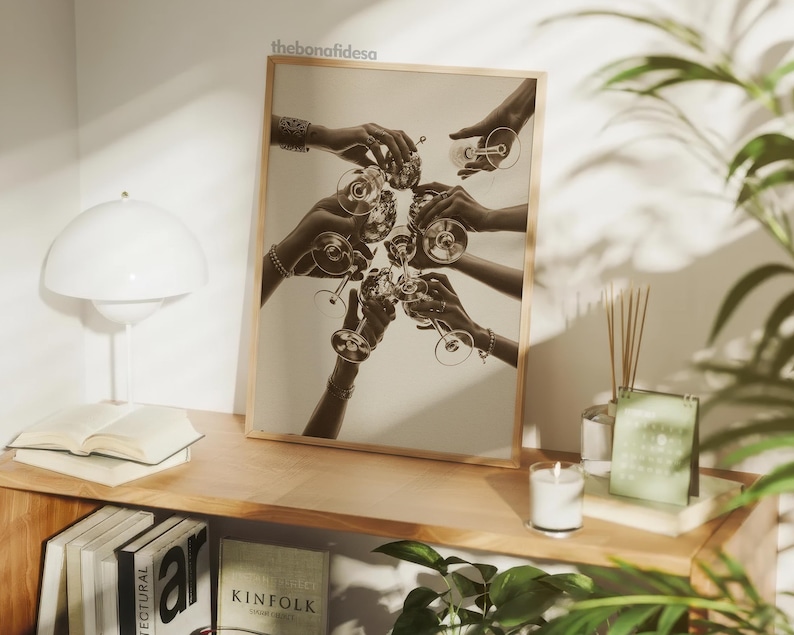 May include: Sepia-toned print of hands toasting with champagne glasses. The image, set in a wooden frame, captures a celebratory moment. The bonafidesa text is visible in the top corner. The print is displayed on a wooden shelf with a lamp, books, and a candle.