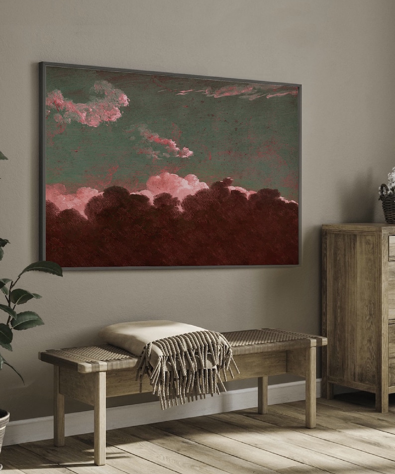 May include: A framed abstract painting with a dark green background and pink clouds. The painting is hanging on a light gray wall above a wooden bench with a beige throw blanket.