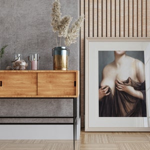 May include: A wooden console table with two sliding doors, a black metal frame, and black metal legs. The table is decorated with a vase of dried flowers, candles, and a decorative object. A framed painting of a woman in a brown dress is hanging on the wall behind the table.