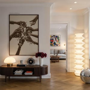 May include: An interior shot featuring a dark wood console table with a lamp, books, and decorative items. A large framed print depicts hands toasting with glasses. A tall, layered lamp and a disco ball are also visible.