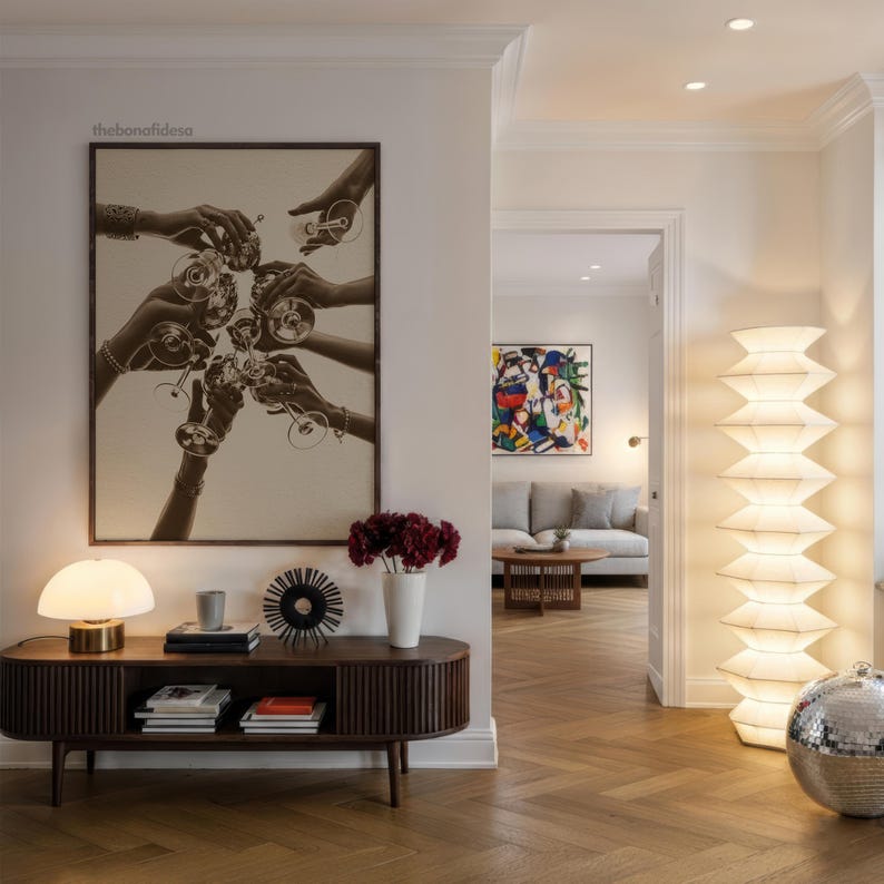 May include: An interior shot featuring a dark wood console table with a lamp, books, and decorative items. A large framed print depicts hands toasting with glasses. A tall, layered lamp and a disco ball are also visible.