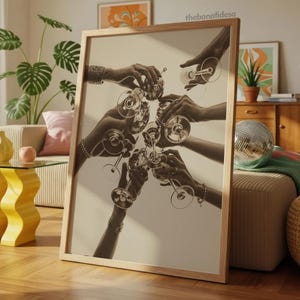 May include: A framed sepia-toned print featuring multiple hands clinking champagne glasses. The image captures a celebratory moment, with the glasses raised in a toast. The artwork is displayed in a light-colored wooden frame, suitable for home decor.