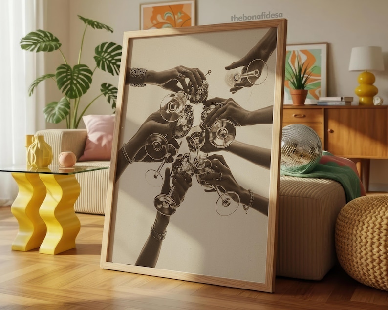 May include: A framed sepia-toned print featuring multiple hands clinking champagne glasses. The image captures a celebratory moment, with the glasses raised in a toast. The artwork is displayed in a light-colored wooden frame, suitable for home decor.