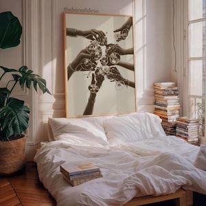 May include: A framed sepia-toned print depicts hands clinking champagne glasses. The artwork hangs above a bed with white linens. A potted plant and stacks of books are visible in the room, creating a cozy, inviting atmosphere.