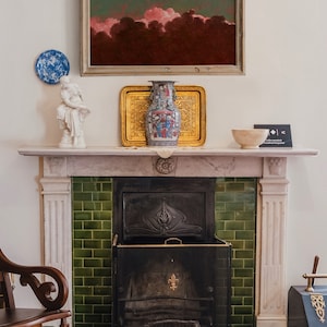 May include: A white marble fireplace with a green tiled surround and a black metal fire grate. A framed painting of a pink and green sky hangs above the fireplace. A white porcelain statue and a blue and white porcelain vase are on the mantel.