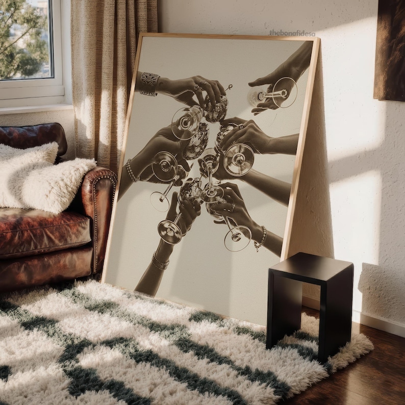 May include: Sepia-toned print of hands toasting with wine glasses. The image is set against a neutral background, with a wooden frame. The print is propped against a wall, near a black side table and a patterned rug.
