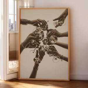 May include: Monochrome print of hands toasting with champagne glasses. The image is framed in a light wood frame. The hands are adorned with bracelets and rings, creating a celebratory atmosphere.