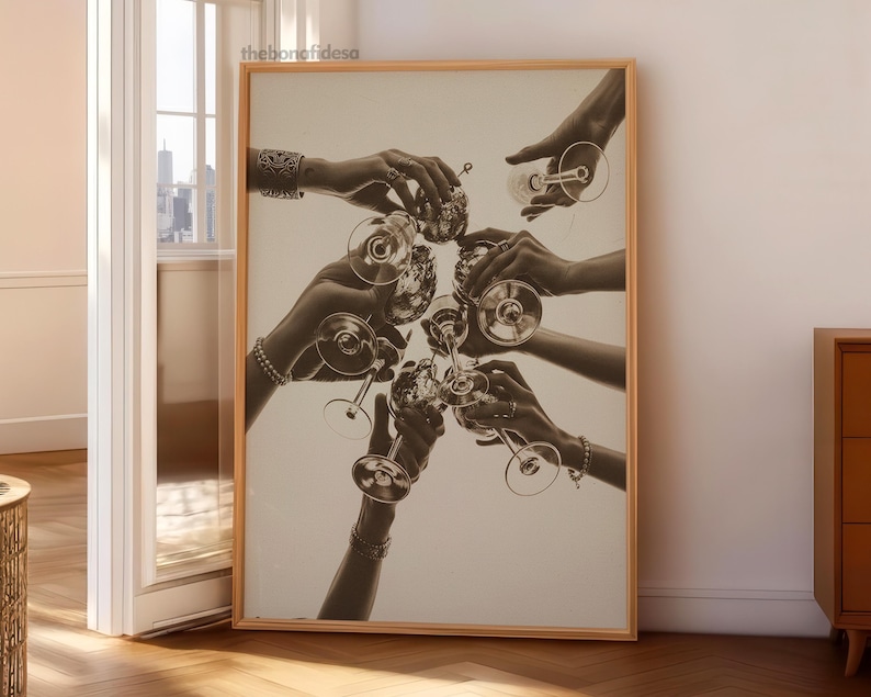 May include: Monochrome print of hands toasting with champagne glasses. The image is framed in a light wood frame. The hands are adorned with bracelets and rings, creating a celebratory atmosphere.