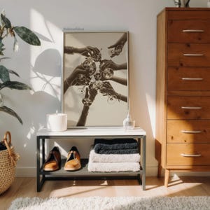 May include: A sepia-toned framed print depicts hands toasting with wine glasses. A white candle, a small glass bottle, and a pair of brown shoes sit on a white and gray table. A stack of towels and a woven basket are also visible.