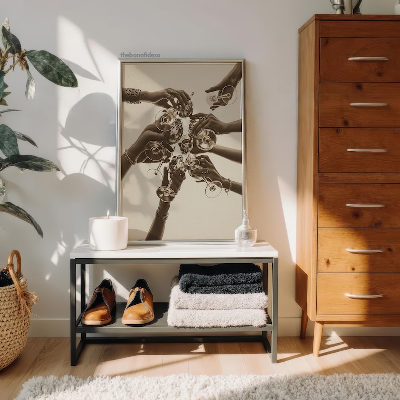May include: A sepia-toned framed print depicts hands toasting with wine glasses. A white candle, a small glass bottle, and a pair of brown shoes sit on a white and gray table. A stack of towels and a woven basket are also visible.