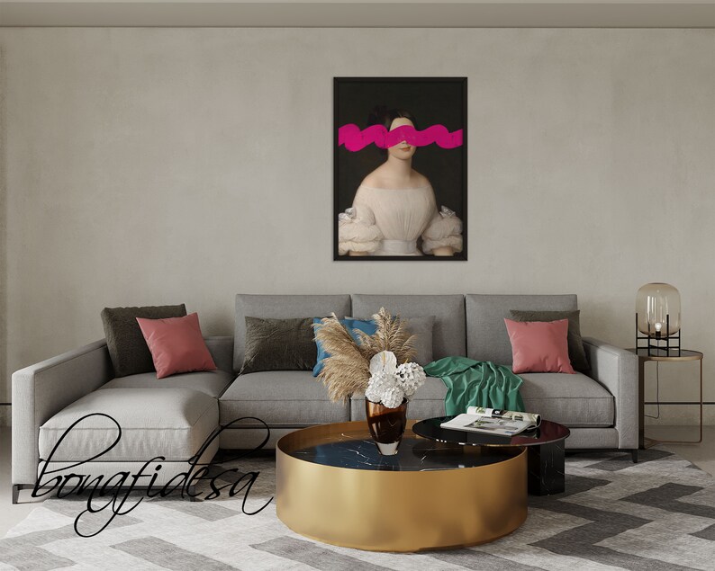 K&ouml;nnte beinhalten: Ein graues Ecksofa mit Kissen und einer Wolldecke. Ein goldener Couchtisch mit einer schwarzen Marmorplatte steht vor dem Sofa. Ein gerahmtes Portr&auml;t einer Frau mit einer pinkfarbenen abstrakten Form, die ihre Augen bedeckt, h&auml;ngt an der Wand hinter dem Sofa.