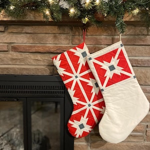 Könnte beinhalten: Zwei rot-weiße Weihnachtsstrümpfe mit geometrischem Muster hängen an einem Kaminsims aus Ziegeln. Die Strümpfe sind mit einem weißen Hintergrund und roten und grauen geometrischen Formen verziert.