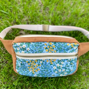 Könnte beinhalten: Braune und floral bedruckte Bauchtasche mit zwei Reißverschlussfächern. Die Bauchtasche hat einen cremefarbenen verstellbaren Riemen und eine goldfarbene Schnalle. Die Vorderseite der Tasche zeigt ein blaues Blumenmuster mit gelben und weißen Blumen.