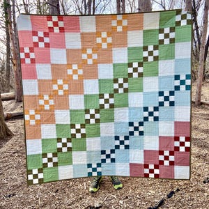 May include: A colourful patchwork quilt with a checkered pattern in shades of red, orange, green, blue, and white. The quilt is made up of squares and is arranged in a diagonal pattern.