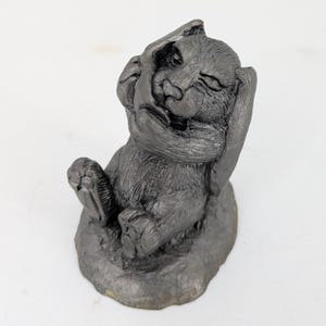 May include: A small, gray, ceramic sculpture of a bear sitting on its haunches with its paws covering its ears. The bear has a playful expression on its face.