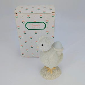 May include: A white ceramic chick figurine with a light blue egg on its back, standing on a beige base. The figurine is next to a box with a floral pattern and the text "Easter 2002".