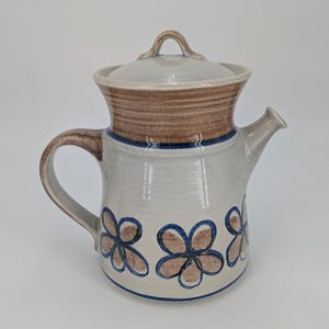 May include: A ceramic teapot with a brown and white floral design. The teapot has a lid and a handle. The teapot is glazed in a light brown color.