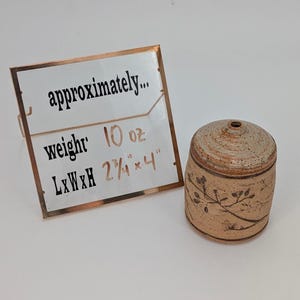 Könnte beinhalten: Ein beigefarbenes Keramikgefäß mit Deckel, verziert mit einem floralen Muster. Das Gefäß ist etwa 7 cm breit und 10 cm hoch und wiegt 283 Gramm. Der Deckel hat ein kleines Loch oben. Der Text auf dem Schild lautet "approximately... weight 10 oz L x W x H 2 3/4" x 4""