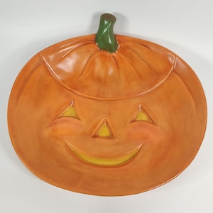 Può includere: Piatto da portata in ceramica arancione a forma di zucca con un viso di zucca intagliato. La zucca ha uno stelo verde e un sorriso giallo.
