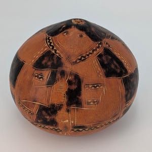 Può includere: Una zucca rotonda e marrone con un disegno intagliato di una persona con un abito a motivi. La zucca presenta sezioni marrone scuro e dettagli incisi, in stile arte popolare.