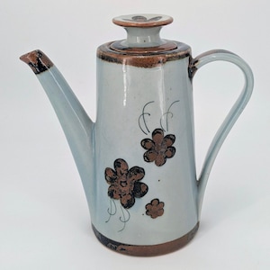 May include: A ceramic teapot with a light blue glaze, featuring brown floral designs. The teapot has a long spout, a curved handle, and a lid with a flower design. The rim and spout are accented with brown. A decorative kitchenware item.