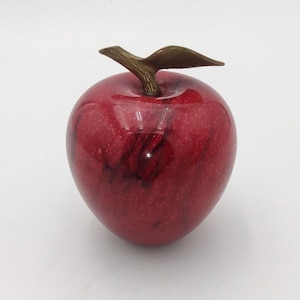 May include: A decorative apple sculpture with a glossy red finish and a bronze-colored stem and leaf. The apple has dark red marbling throughout. This piece is a decorative accent for a home or office.