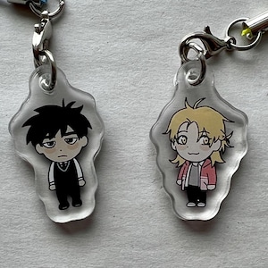 May include: Two anime-style character charms. One charm features a character with black hair and a black vest. The other charm has a character with blonde hair and a red jacket. Each charm has a lanyard.