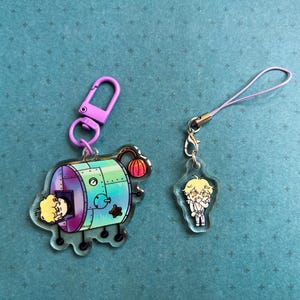 Peut inclure: Deux breloques en acrylique transparent. L'une est une machine violette et sarcelle avec un personnage de dessin animé à l'intérieur et une boule rouge sur le dessus. L'autre est un petit charme d'un personnage de dessin animé avec un cordon rose.