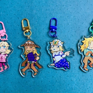 Può includere: Quattro portachiavi in acrilico con personaggi in stile anime con vari abiti e pose. Ogni portachiavi ha un fermaglio di colore diverso: rosa, oro, blu e verde. I personaggi hanno i capelli biondi e sono su uno sfondo blu.