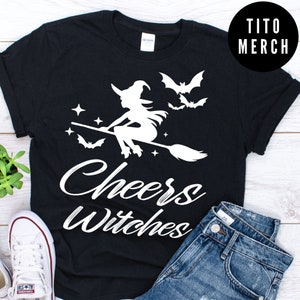 May include: Black t-shirt with a white silhouette of a witch flying on a broomstick with bats and stars. The text "Cheers Witches" is written in a white cursive font.