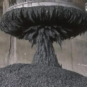 Pode incluir: Uma grande máquina industrial dispensando uma substância escura e granular. O material forma uma forma de cogumelo ao cair sobre uma pilha. A máquina é metálica e o fundo é cinza neutro.