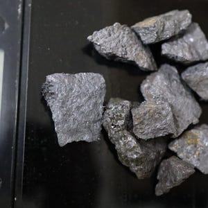 May include: A collection of dark grey, jagged mineral rocks resting on a black digital scale. The scale display shows a weight of 3.15 grams. The rocks have a rough, crystalline texture with visible metallic flecks across their uneven surfaces.