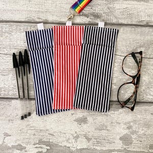 May include: Three striped pen cases in navy, red, and black, with a pair of eyeglasses and several black pens. The cases have a vertical stripe pattern. A rainbow lanyard is attached to the top of the cases.